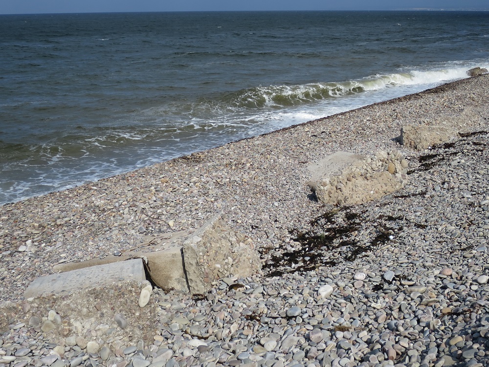 Tank traps that have been buried by the shingle, then exposed again, and have now fallen and are being destroyed.